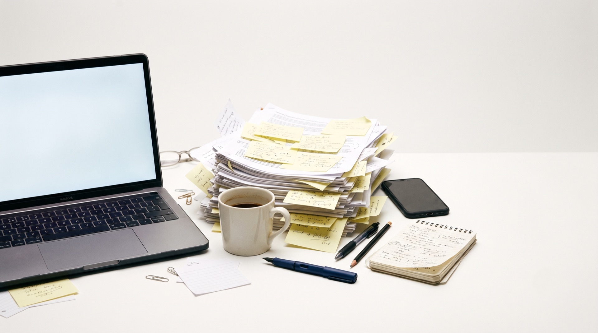 A white desk with an open laptop, a thick stack of printed program lists covered in sticky notes, and a cold cup of coffee.